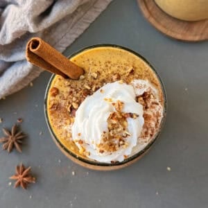 Pumpkin pie protein shake in a glass with a cinnamon stick and topped with whip cream, crushed pecans, and cinnamon powder.
