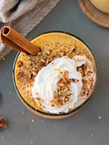 Pumpkin pie protein shake in a glass with a cinnamon stick and topped with whip cream, crushed pecans, and cinnamon powder.