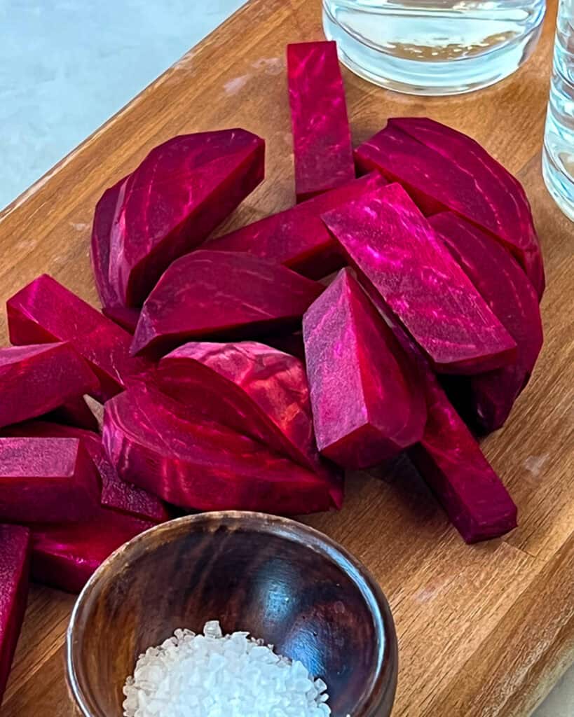 Sliced raw beets with a small bowl of slat on a flat wooden cutting board.