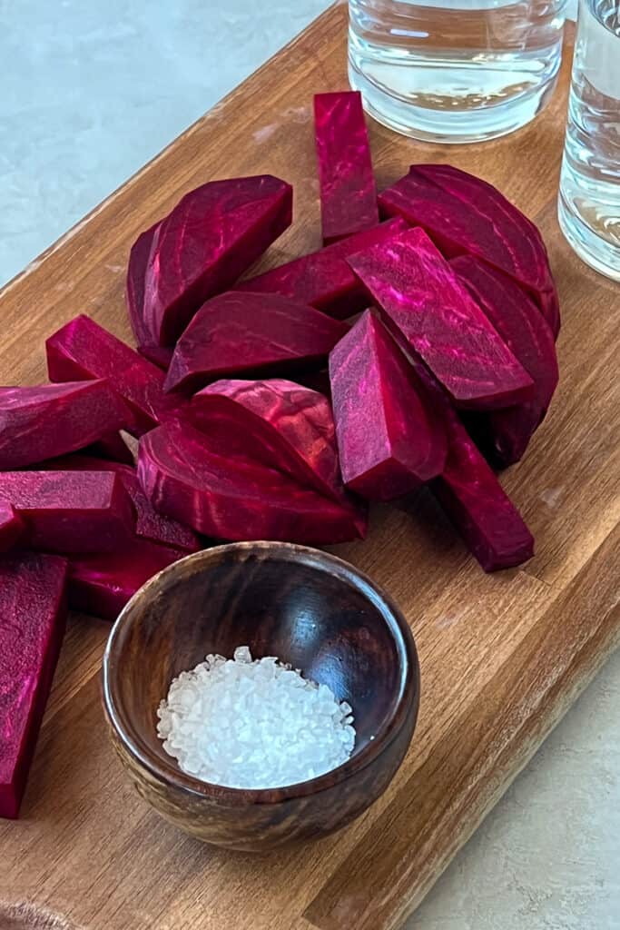 Sliced raw beets with a small bowl of slat on a flat wooden cutting board.