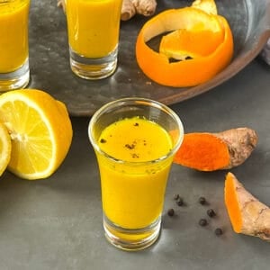 Ginger turmeric shot in a 2 ounce glass next to fresh whole ingredients.